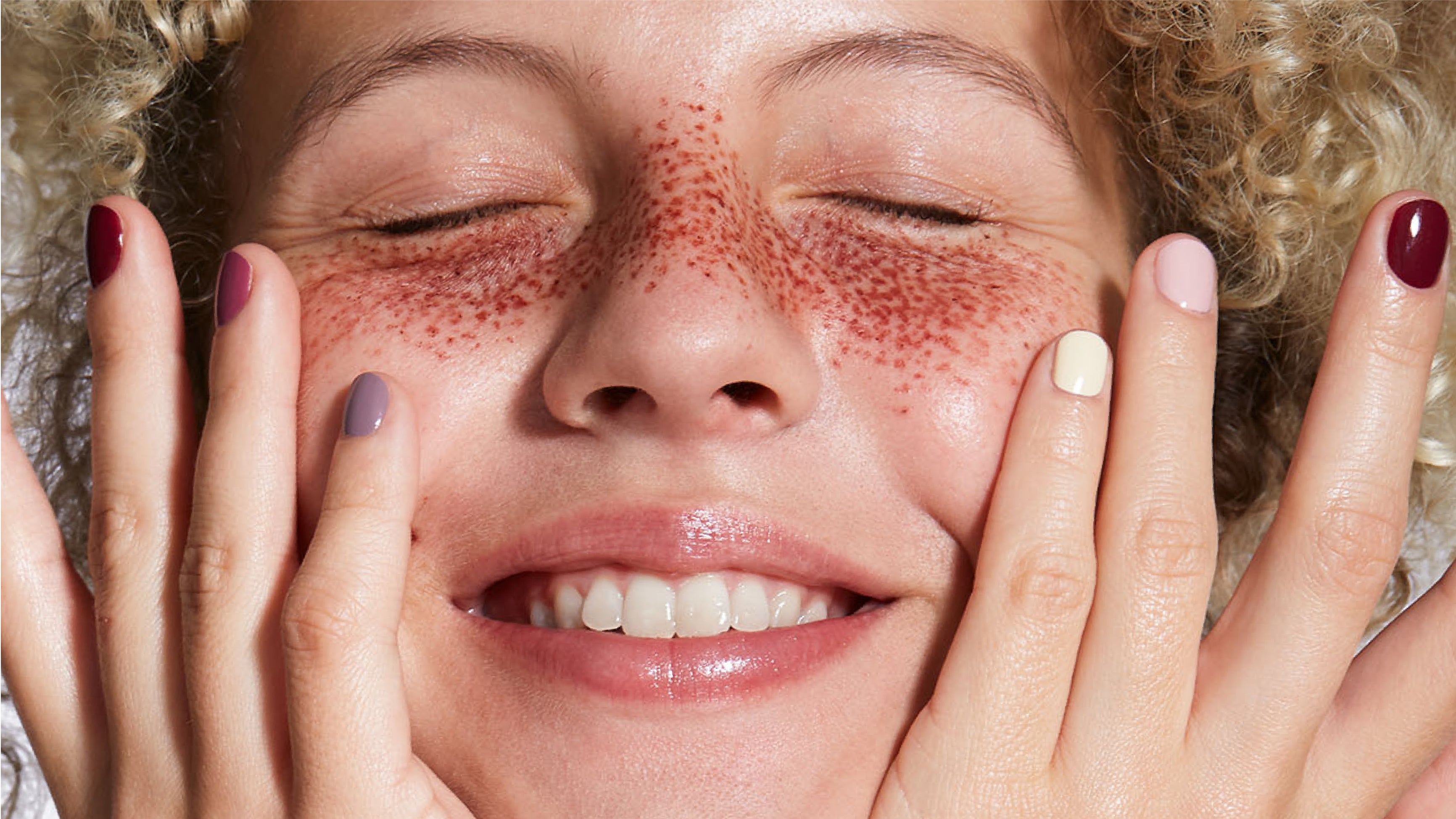 Eine Person mit geschlossenen Augen und sichtbaren Sommersprossen lächelt und berührt ihr Gesicht mit beiden Händen; jeder Fingernagel ist in einer anderen Farbe bemalt.