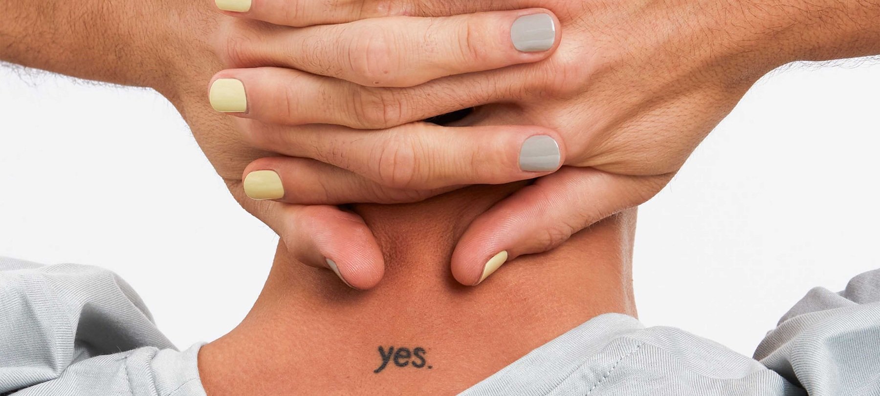 Nahaufnahme des Nackens einer Person mit einem kleinen Ja-Tattoo und Händen mit pastellgelbem und grauem Nagellack, die auf ihrem Kopf ruhen.
