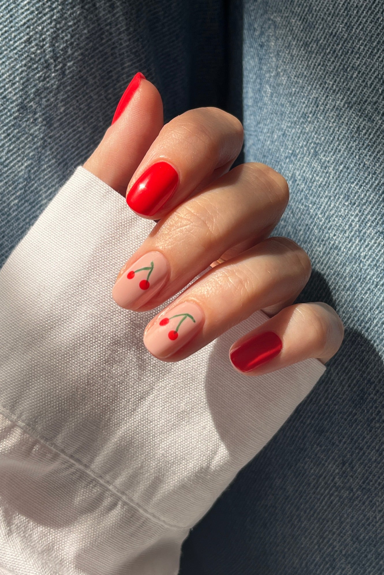 Eine Hand mit rot lackierten Nägeln unter Verwendung des Nailart-Sets für einen Ober- und Unterlack und Kirschdesigns auf zwei nackten Nägeln. Die Person trägt ein weißes langärmeliges Hemd und eine blaue Jeans.