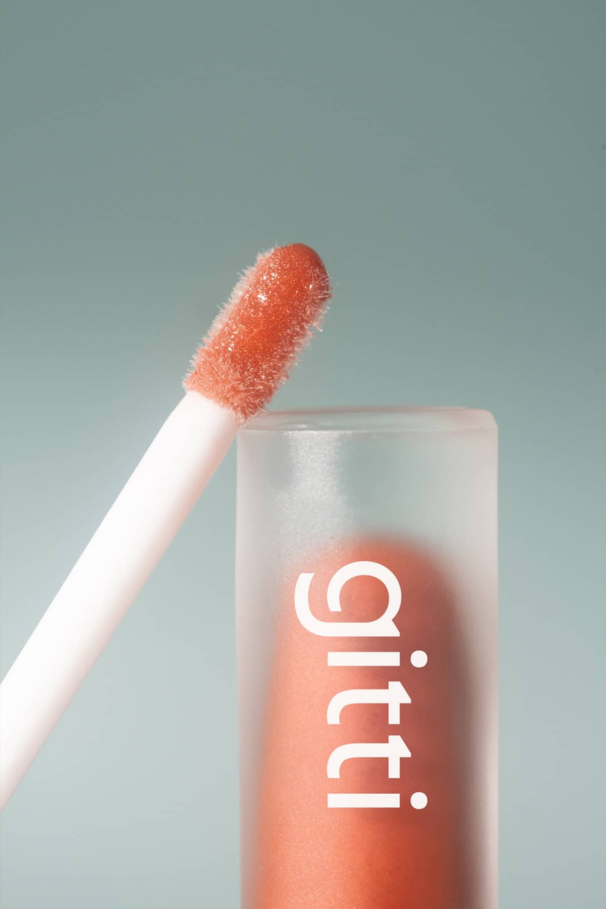 Eine Nahaufnahme des Lip Glaze no. 04 Applikator mit Nude-Ton und Hochglanz-Finish, neben einer mattierten Tube mit der Aufschrift gitti auf hellblauem Hintergrund.