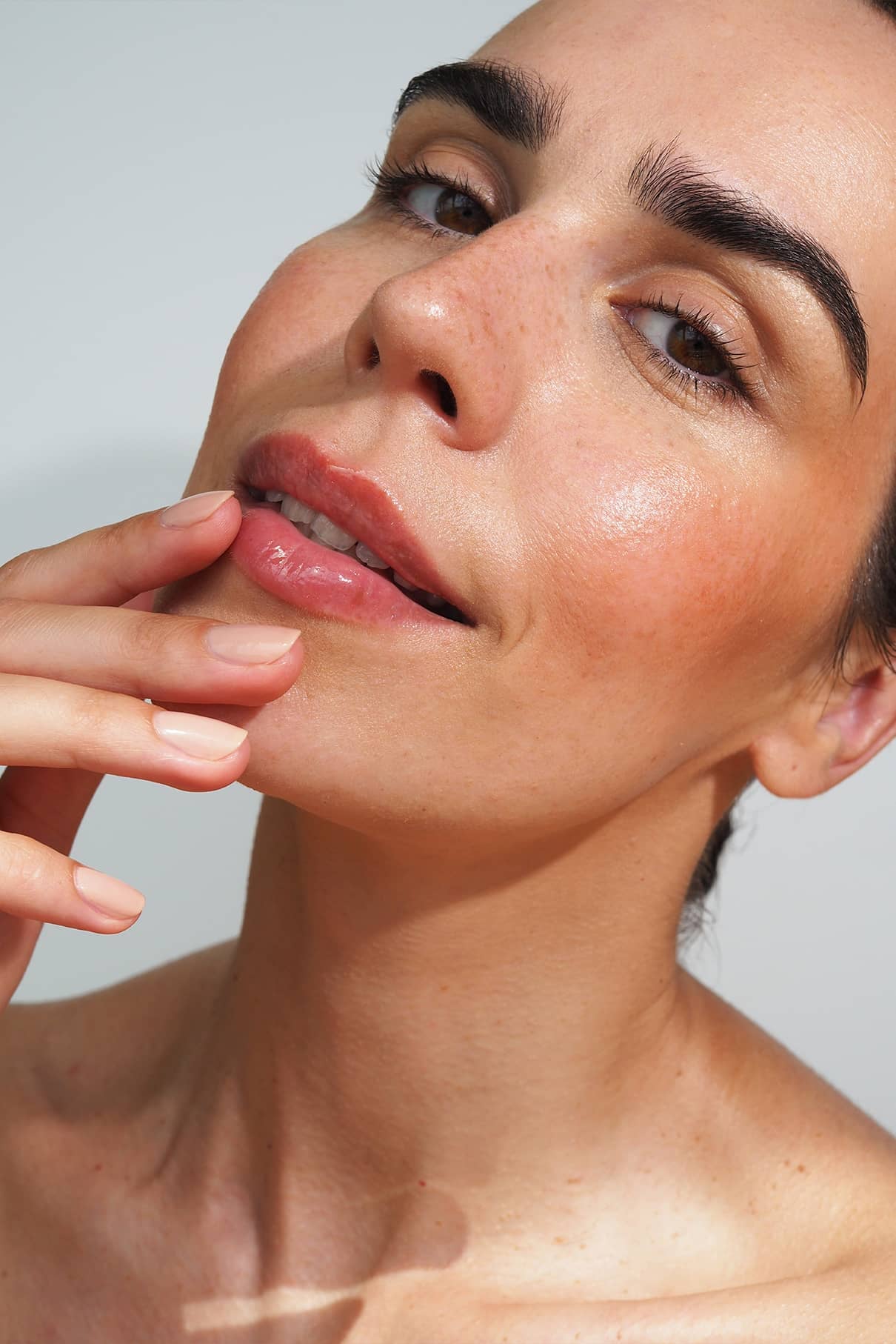 Eine Person mit klarer, strahlender Haut und natürlichem Make-up berührt sanft ihre Lippen und hebt das hochglänzende Finish von Lip Glaze no. 04 hervorhebt, alles vor einem schlichten Hintergrund.