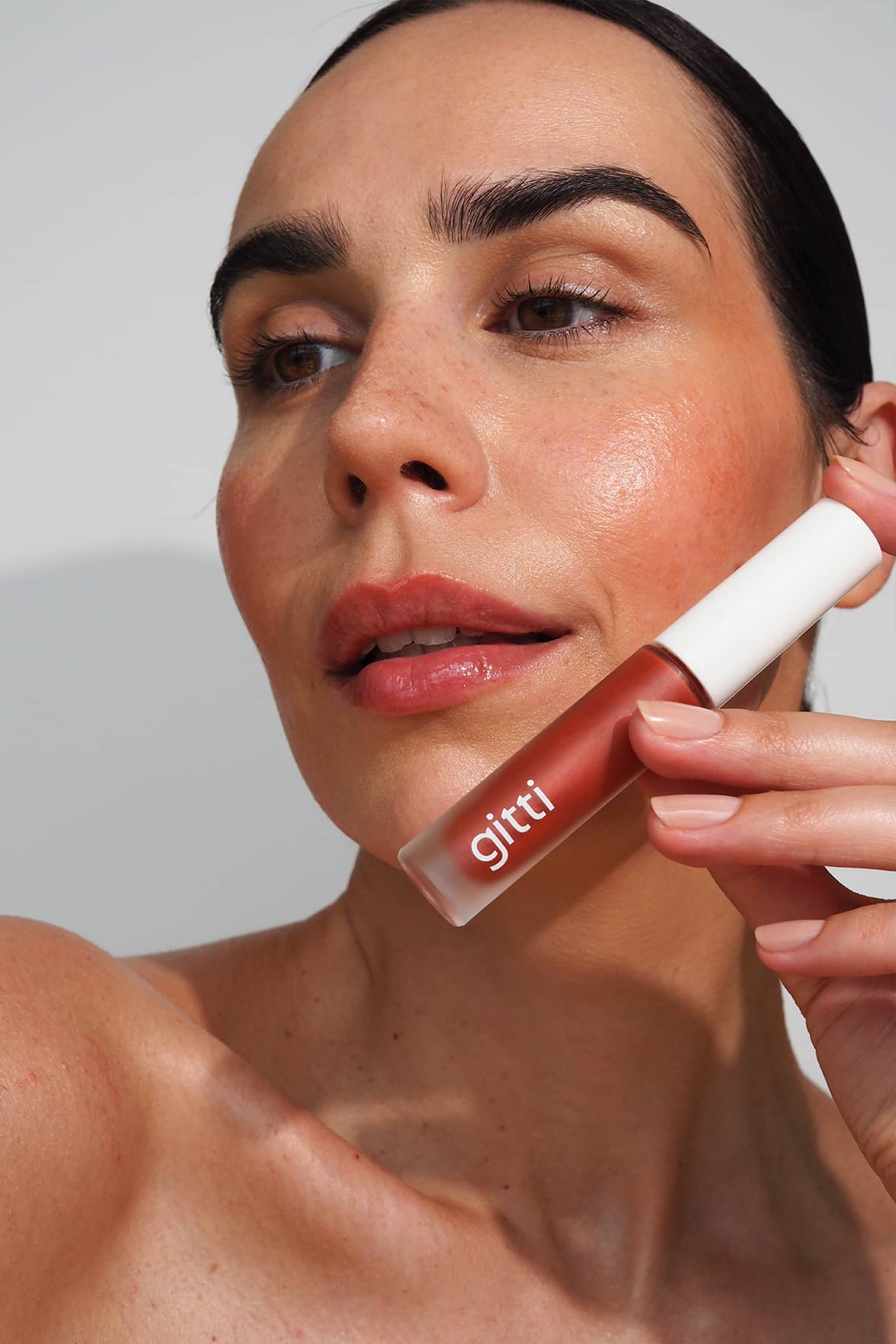 Eine Frau mit taufrischer Haut hält eine Tube Lip Glaze no. 04 nahe an ihr Gesicht und zeigt den Applikator und das hochglänzende Finish auf ihren Lippen.