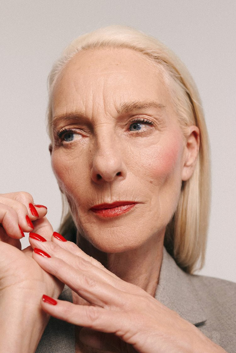 Ältere Frau mit glattem, schulterlangem blondem Haar trägt einen grauen Blazer und Velvet Cheeks no. 01 mousse-blush. Sie schaut mit den Händen neben ihrem Gesicht, rot lackierten Nägeln und silikonfreiem Make-up zur Seite.