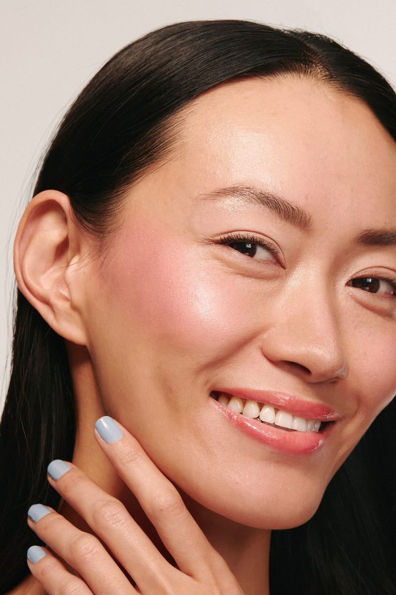 Eine lächelnde Frau mit glattem schwarzen Haar und klarer, strahlender Haut trägt Rouge und hellblauen Nagellack aus dem Summer Glow Set. Ihre Hand ruht neben ihrem Gesicht, während mit Mascara verstärkte Wimpern ihren strahlenden Blick vervollständigen.