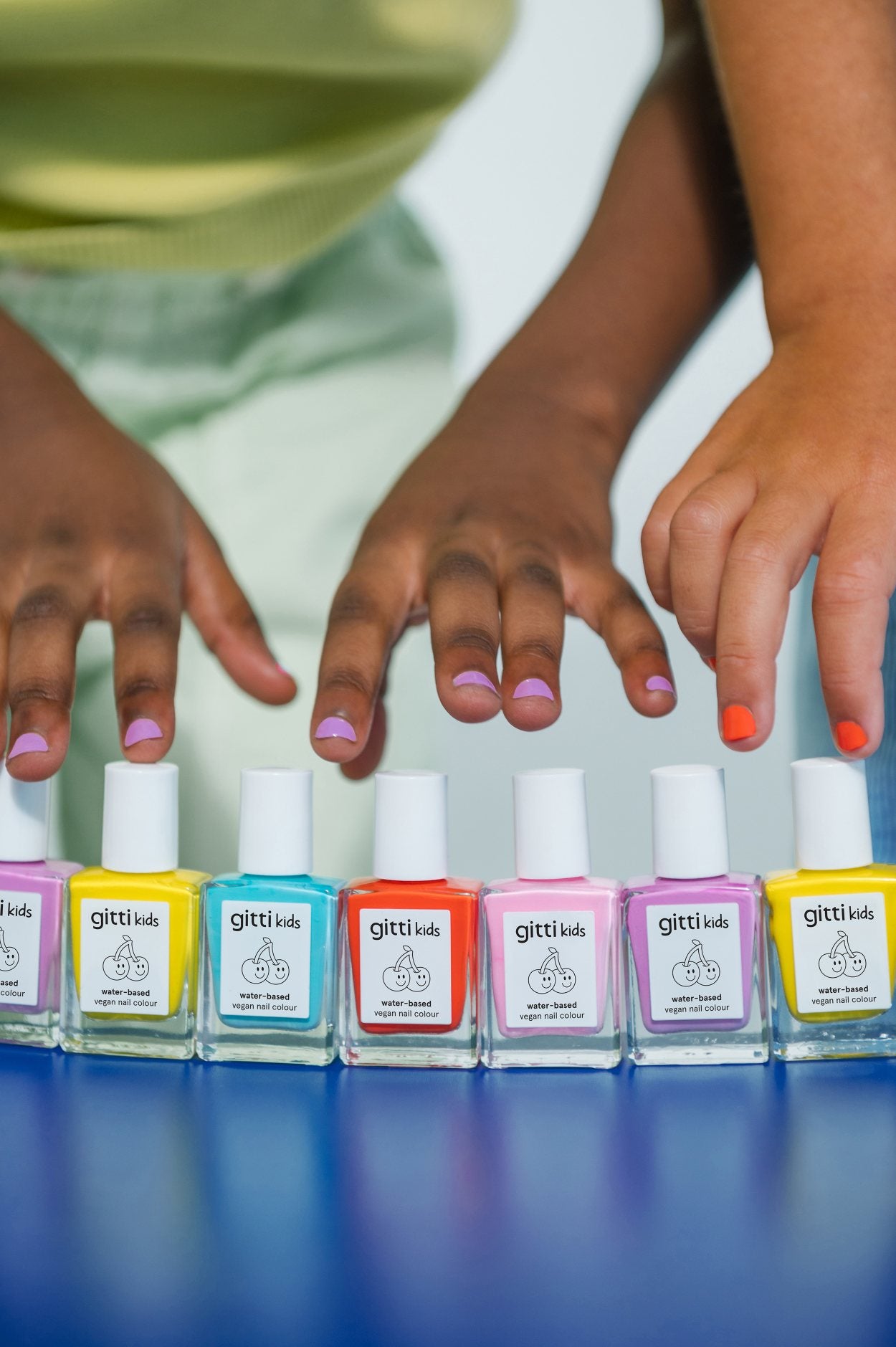Zwei Hände mit bunt lackierten Nägeln stehen über einer Reihe von sieben Fläschchen mit Kids Duo Nagellack in verschiedenen leuchtenden Farben auf einer blauen Fläche.