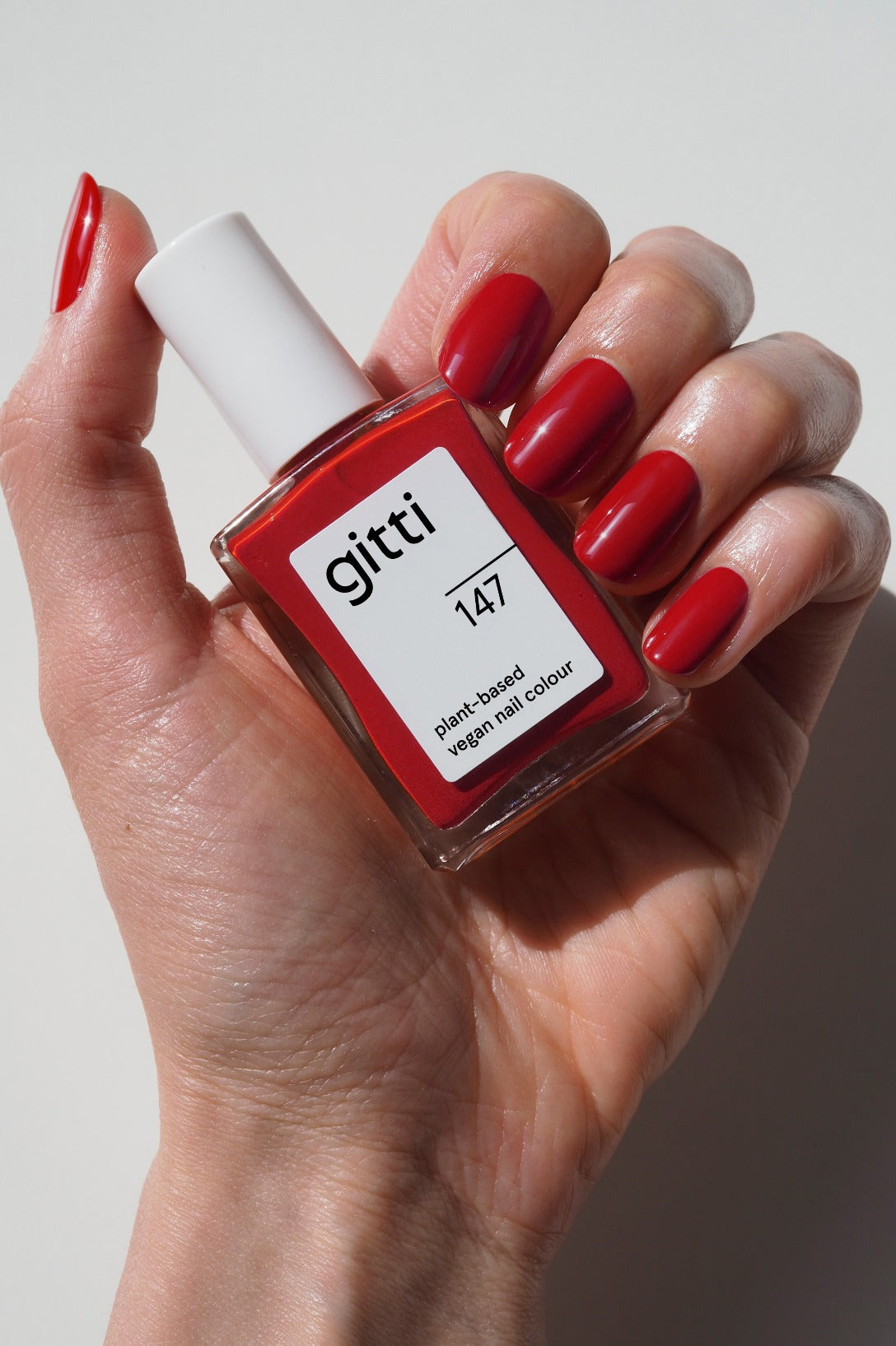 Eine Hand mit rot lackierten Nägeln hält ein Fläschchen mit gitti no. 147 pflanzliche vegane Nagelfarbe in klassischem Rot vor einem schlichten weißen Hintergrund.