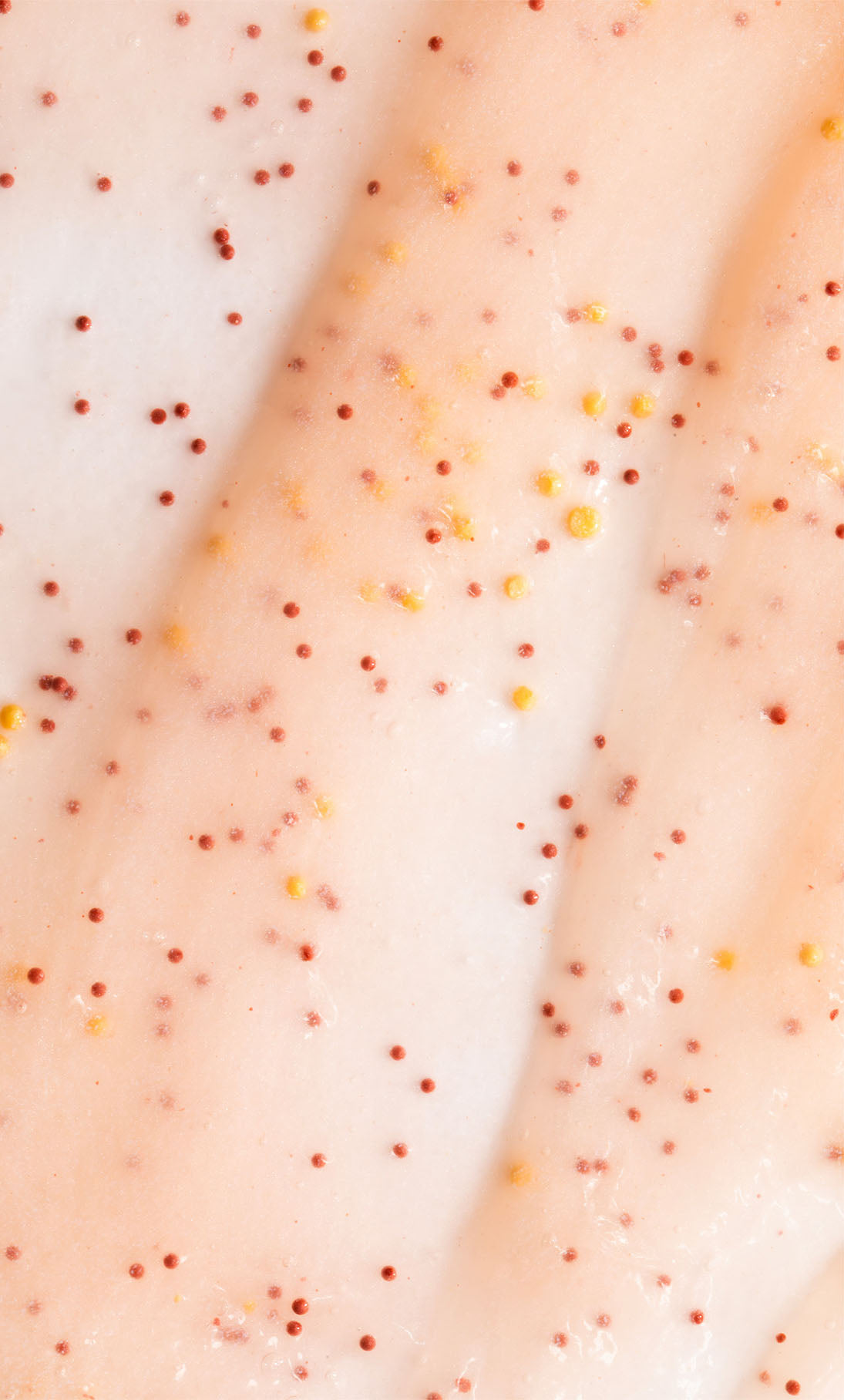 Nahaufnahme des AHA Jelly Hand Exfoliator: eine cremige, blassrosa Formel mit roten und gelben Peelingperlen und einem gleichmäßig verteilten AHA-Komplex.