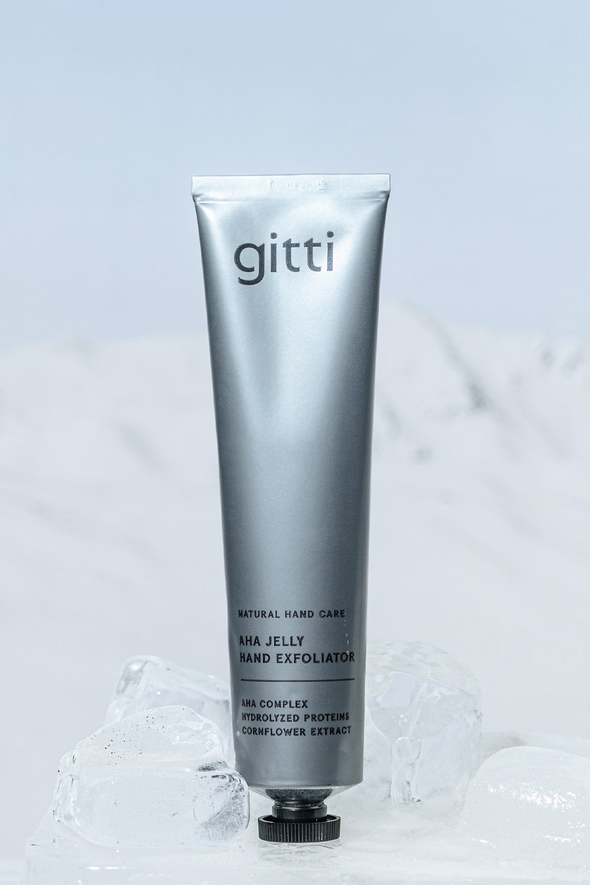 Eine silberne Tube des AHA Jelly Hand Exfoliator steht aufrecht auf klaren Eisblöcken vor einem hellen, strukturierten Hintergrund.