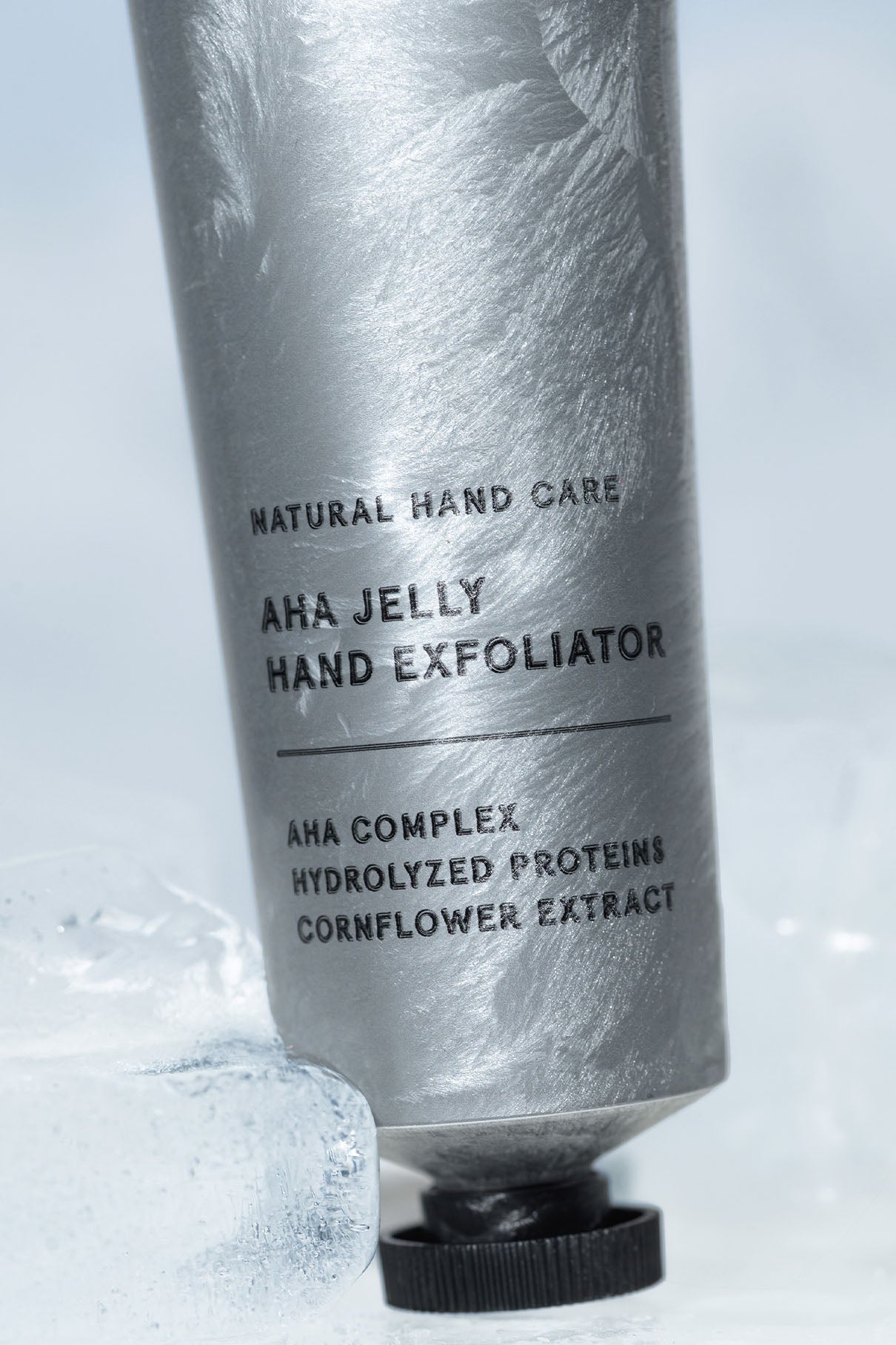 Eine silberne Tube mit der Aufschrift AHA Jelly Hand Exfoliator steht aufrecht auf Eis und enthält einen Text über den AHA-Komplex, hydrolysierte Proteine und beruhigenden Kornblumenextrakt.