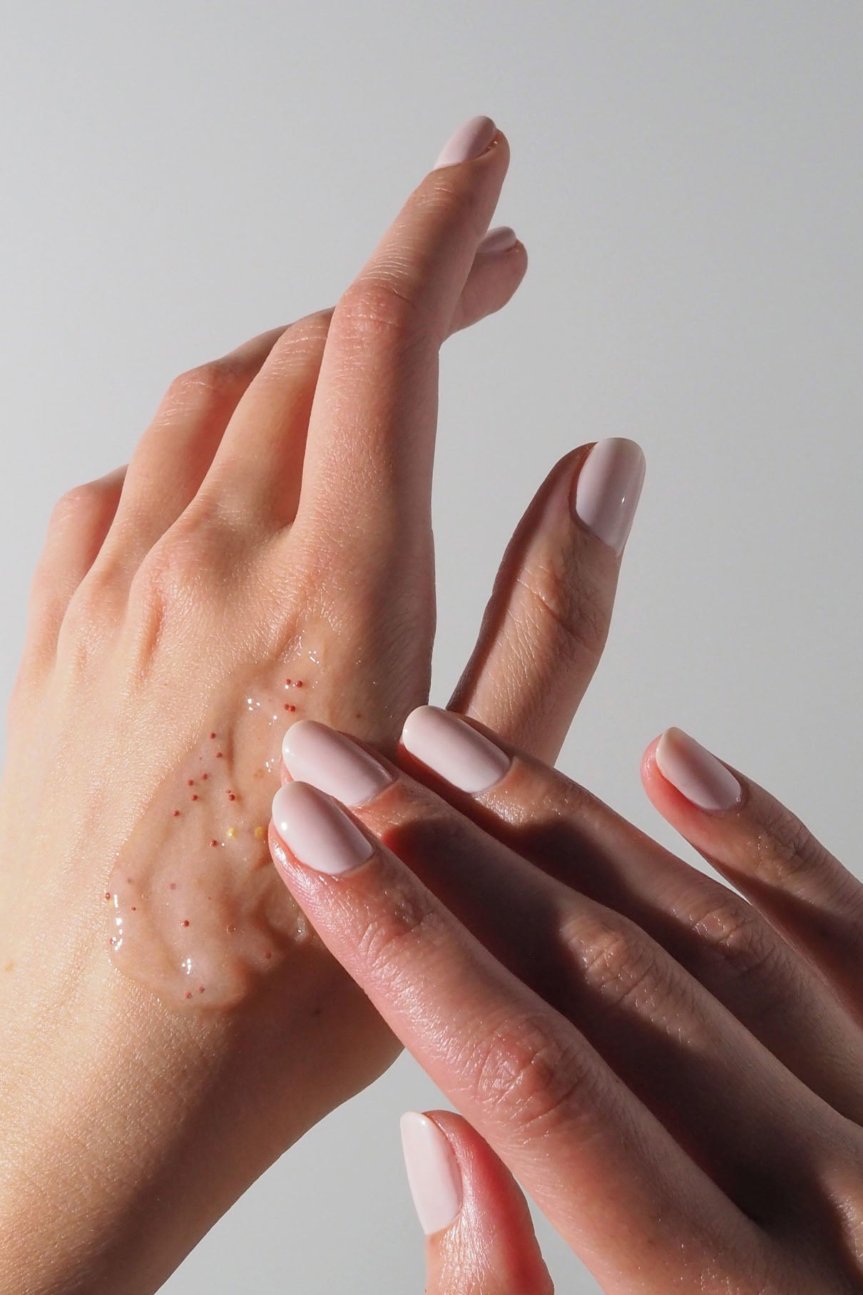 Eine Person trägt Silky Hands Duo, ein durchscheinendes Handpeeling-Gel mit kleinen roten Partikeln, auf den Handrücken auf und hebt damit ihre gepflegten blassrosa Nägel vor einem unifarbenen Hintergrund hervor.
