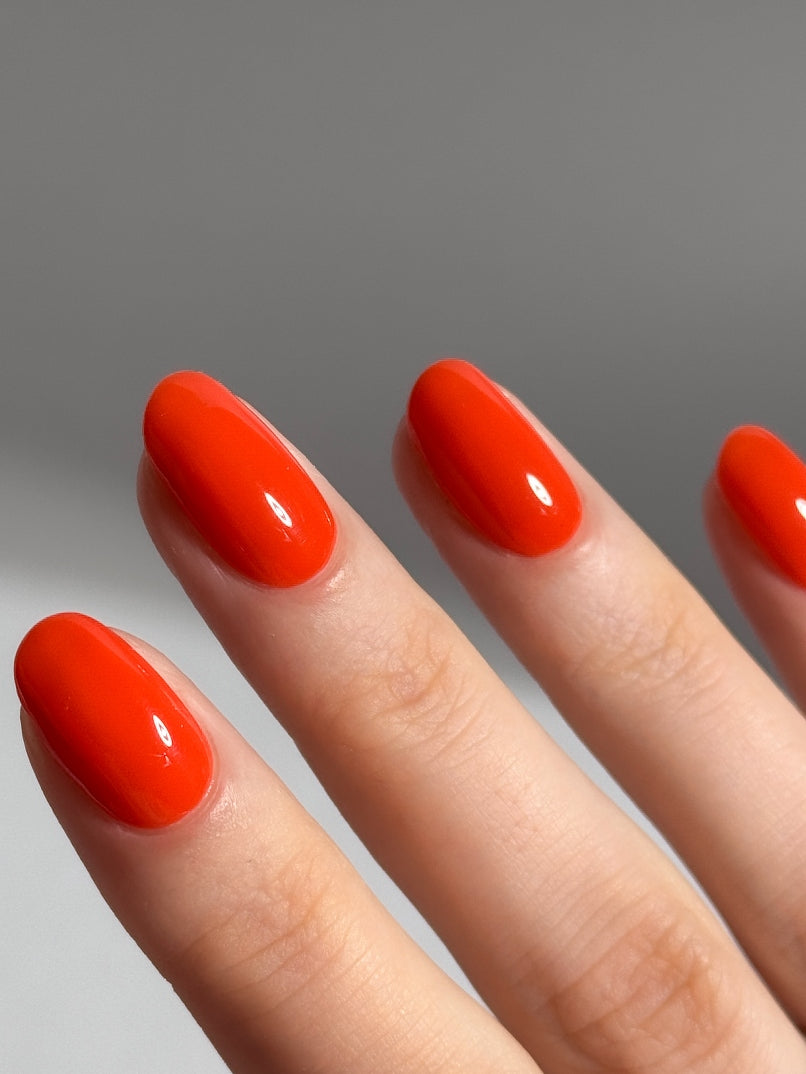 Eine Nahaufnahme einer Hand mit sauber manikürten Nägeln, die mit glänzendem, leuchtend rot-orangem Lack lackiert und mit dem plantPRO Top Coat veredelt wurden, zeigt einen gelartigen Glanz vor einem schlichten Hintergrund.