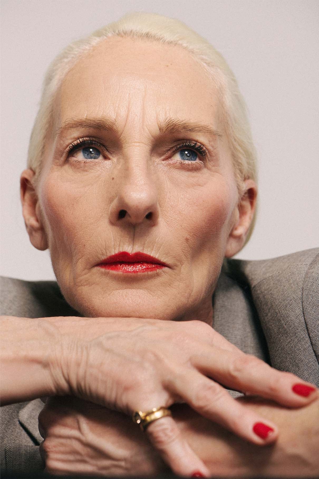 Nahaufnahme einer älteren Frau mit heller Haut und blauen Augen, die Lip Glaze no. 02 in hochglänzendem Finish und stützt ihre Hände unter dem Kinn ab. Sie ist mit einem grauen Blazer und einem goldenen Ring bekleidet.