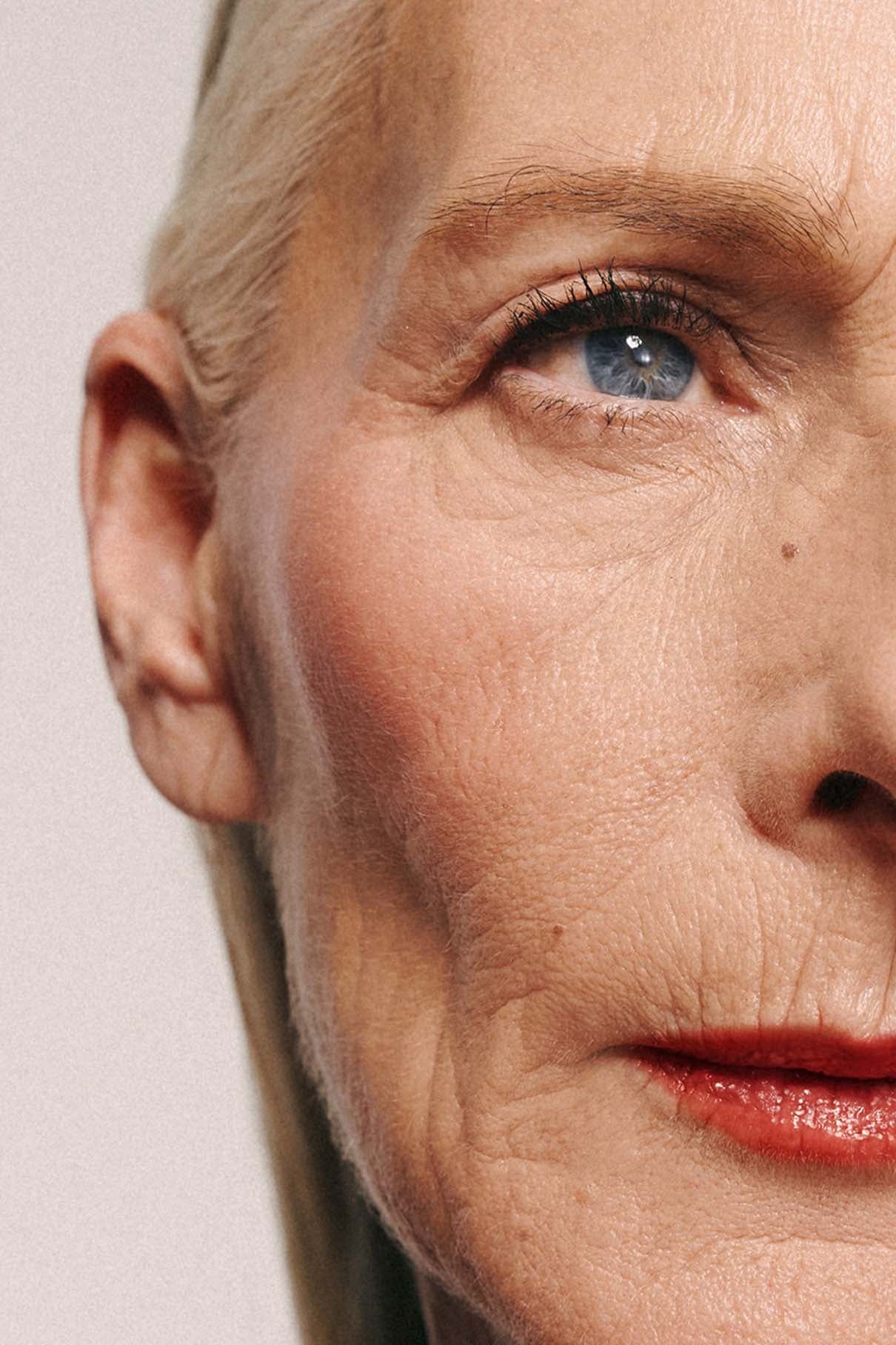Nahaufnahme des Gesichts einer älteren Frau, die ein blaues Auge, helle Haut mit Falten, Velvet Cheeks no. 01 auf den Wangen, roten Lippenstift und blondes, nach hinten gezogenes Haar.