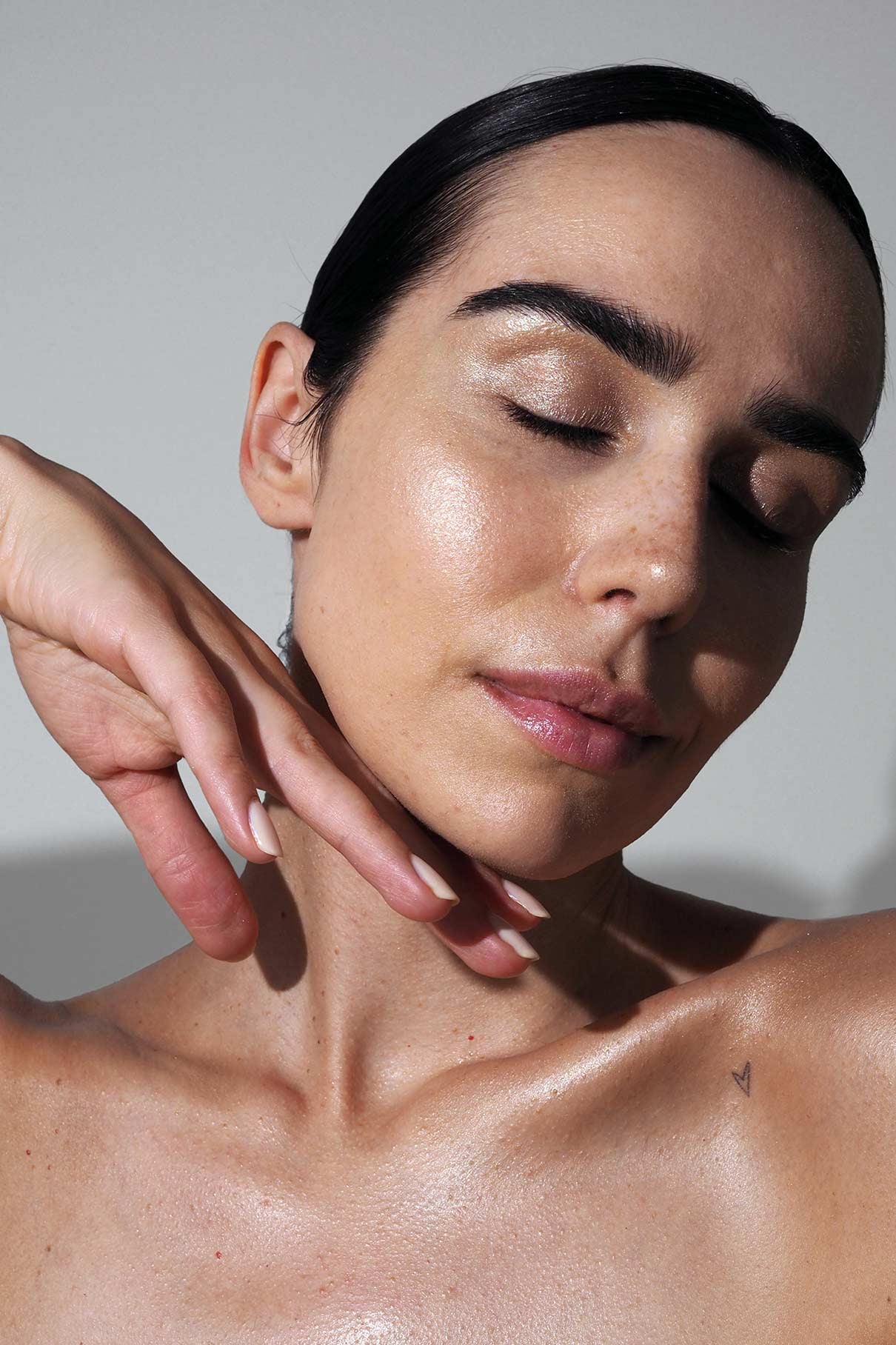 Eine Person mit dunklen Haaren und gepflegten Augenbrauen schließt die Augen, neigt den Kopf und präsentiert eine strahlende, taufrische Haut, die sie mit dem Summer Glow Set, üppiger Wimperntusche und einem kleinen Herz-Tattoo auf der Schulter erzielt hat.