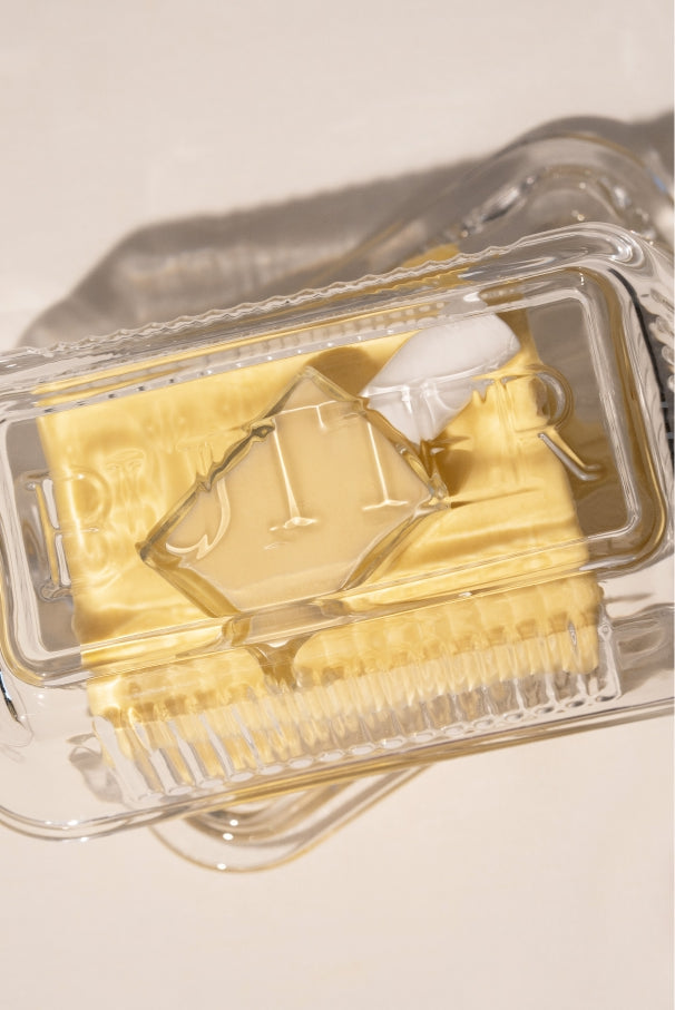 Eine Butterstange in einer Butterdose aus Klarglas mit Deckel, von oben auf neutralem Hintergrund fotografiert, zeigt den Farbton gitti no. 032 mit seinen sanften gelben und cremigen weiß-gelben Farbtönen.