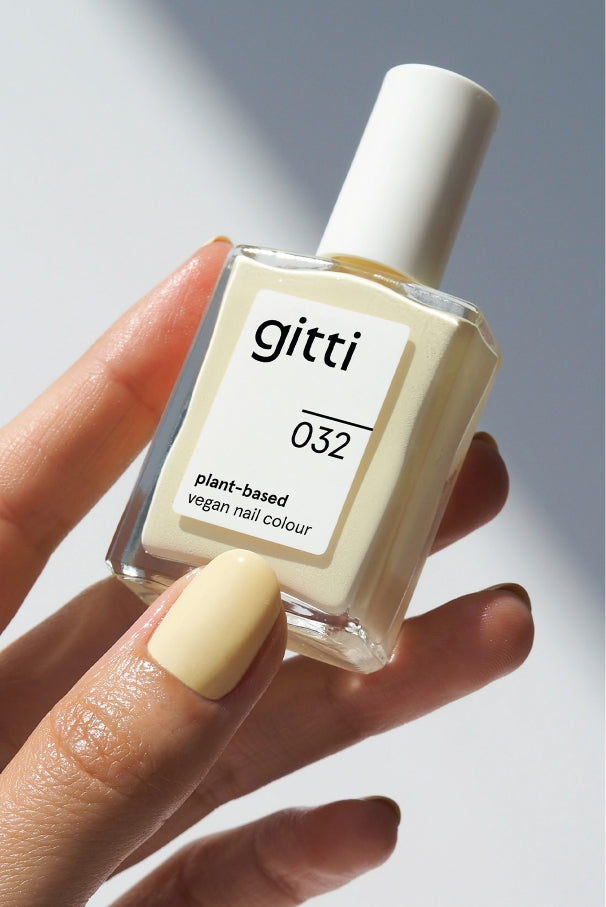 Eine Hand mit blassgelb lackierten Nägeln hält eine Flasche gitti no. 032, einer pflanzlichen, veganen Nagelfarbe in einem zarten, butterweichen Weißgelb.