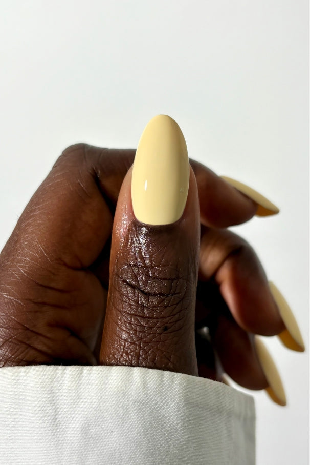 Nahaufnahme einer Hand mit mandelförmigen Nägeln, die in gitti no. 032, einem zarten Gelbton, der sich sanft von einem hellen langärmeligen Hemd abhebt.