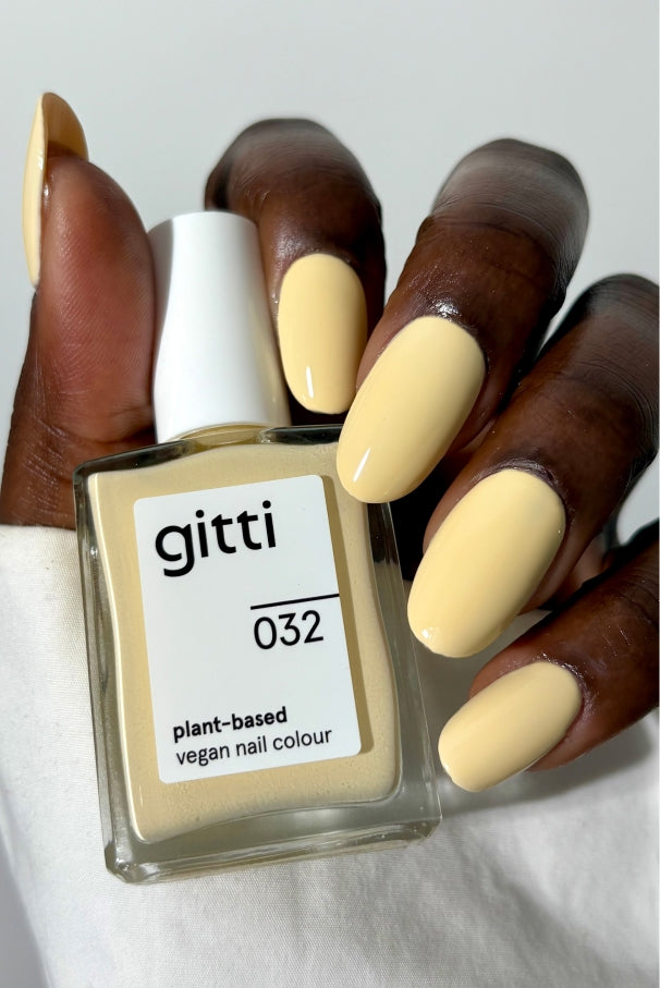 Eine Hand mit blassgelben Nägeln hält eine Flasche gitti no. 032, einer pflanzlichen, veganen Nagelfarbe in sanftem Gelb, die einen weichen und eleganten Farbton aufweist.