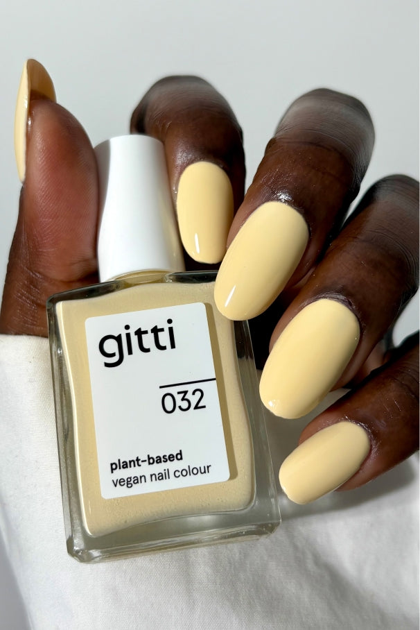Eine Hand mit blassgelben Nägeln hält eine Flasche gitti no. 032, einer pflanzlichen, veganen Nagelfarbe in sanftem Gelb, die einen weichen und eleganten Farbton aufweist.