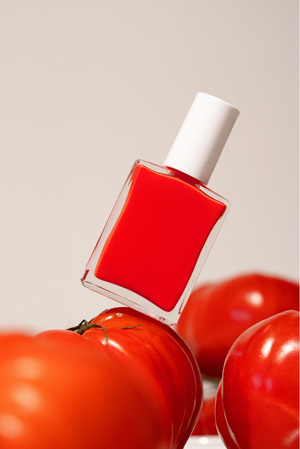 Eine Flasche gitti no. 033, ein roter Nagellack mit weißer Kappe, steht inmitten reifer roter Tomaten vor einem schlichten Hintergrund.