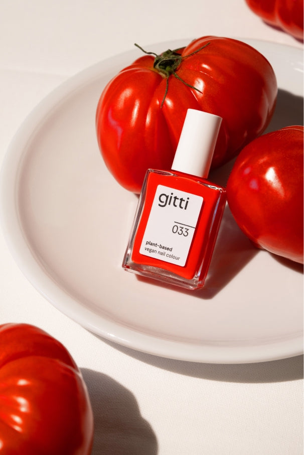 Eine Flasche gitti no. 033 Nagellack in intensivem Tomatenrot wird auf einem weißen Teller platziert, begleitet von drei großen roten Tomaten.