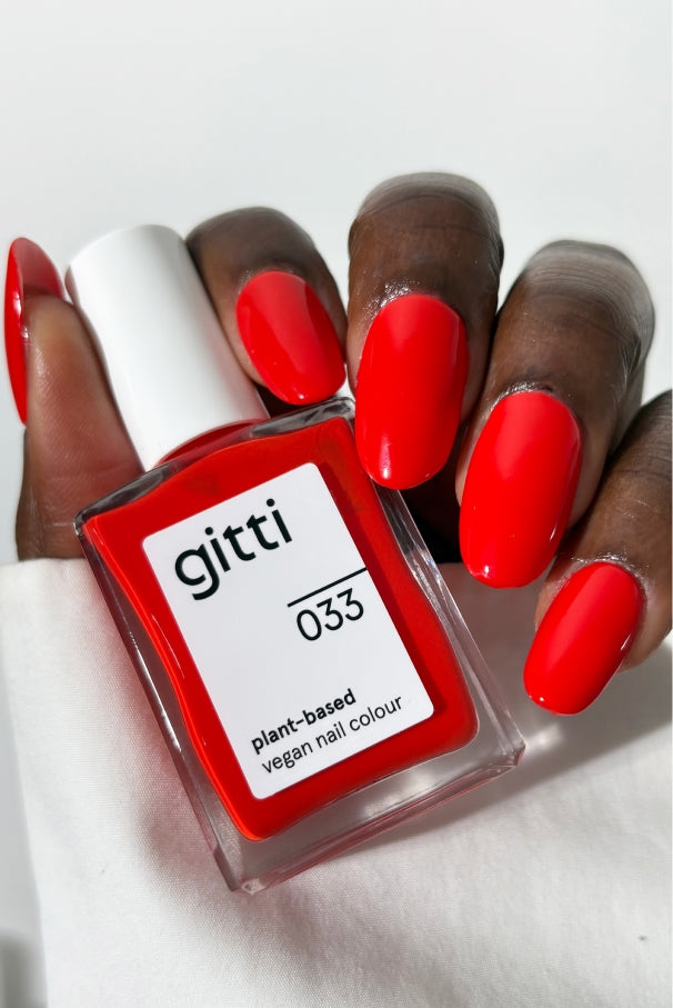Eine Hand mit leuchtend rot lackierten Nägeln hält ein Fläschchen mit gitti no. 033, einer pflanzlichen, veganen Nagelfarbe in einem intensiven Tomatenrot.