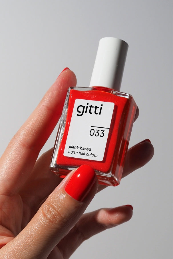 Eine Hand mit rot lackierten Nägeln hält eine quadratische Flasche gitti no. 033 pflanzlicher veganer Nagelfarbe in leuchtendem Tomatenrot vor einem schlichten Hintergrund.