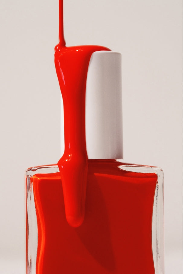 Ein Fläschchen mit gitti no. 033 Nagellack in intensivem Tomatenrot, mit leuchtend rotem Lack, der über die weiße Kappe fließt und heruntertropft, vor einem neutralen Hintergrund.
