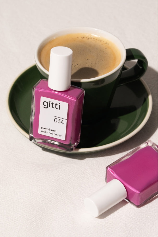 Eine Flasche gitti no. 034 veganer Nagellack steht vor einer Tasse Kaffee auf einer grünen Untertasse, eine weitere gitti no. 034-Flasche liegt daneben auf der weißen Fläche.