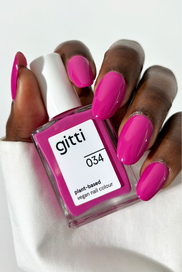 Eine Hand mit langen, rosa lackierten Nägeln hält eine Flasche gitti no. 034 pflanzlicher, veganer Nagellack in elektrisierendem Fuchsia auf weißem Hintergrund.