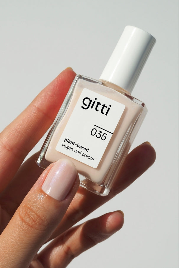 Eine Hand mit hellrosa Nagellack hält eine Flasche gitti no. 035, eine pflanzliche, vegane Nagelfarbe in einem halbtransparenten, sanften Rosé-Ton, vor einem schlichten Hintergrund.