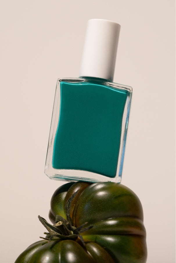 Eine Flasche von gitti no. 036, ein blaugrüner Nagellack mit weißer Kappe, wird auf einer dunkelgrünen Erbstücktomate balanciert und hebt ihren tiefen Grün-Blau-Ton vor einem neutralen Hintergrund hervor.
