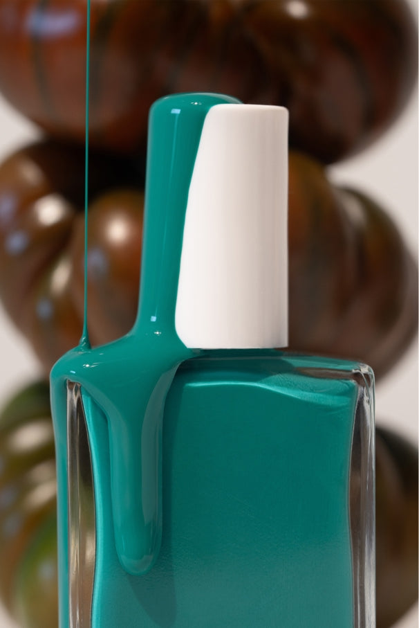 Eine Flasche gitti no. 036, ein blaugrüner Nagellack in einem mediterranen Grün-Blau-Ton, bei dem der Lack an der weißen Kappe heruntertropft; im Hintergrund sind verschwommene braune Tomaten zu sehen.