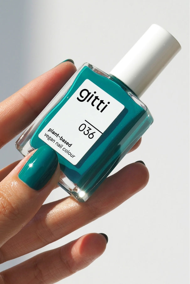 Eine Hand mit blaugrün lackierten Nägeln hält eine Flasche gitti no. 036, einer veganen, pflanzlichen Nagelfarbe in tiefem Grün-Blau, inspiriert von mediterranen Farbtönen, vor einem weißen Hintergrund.