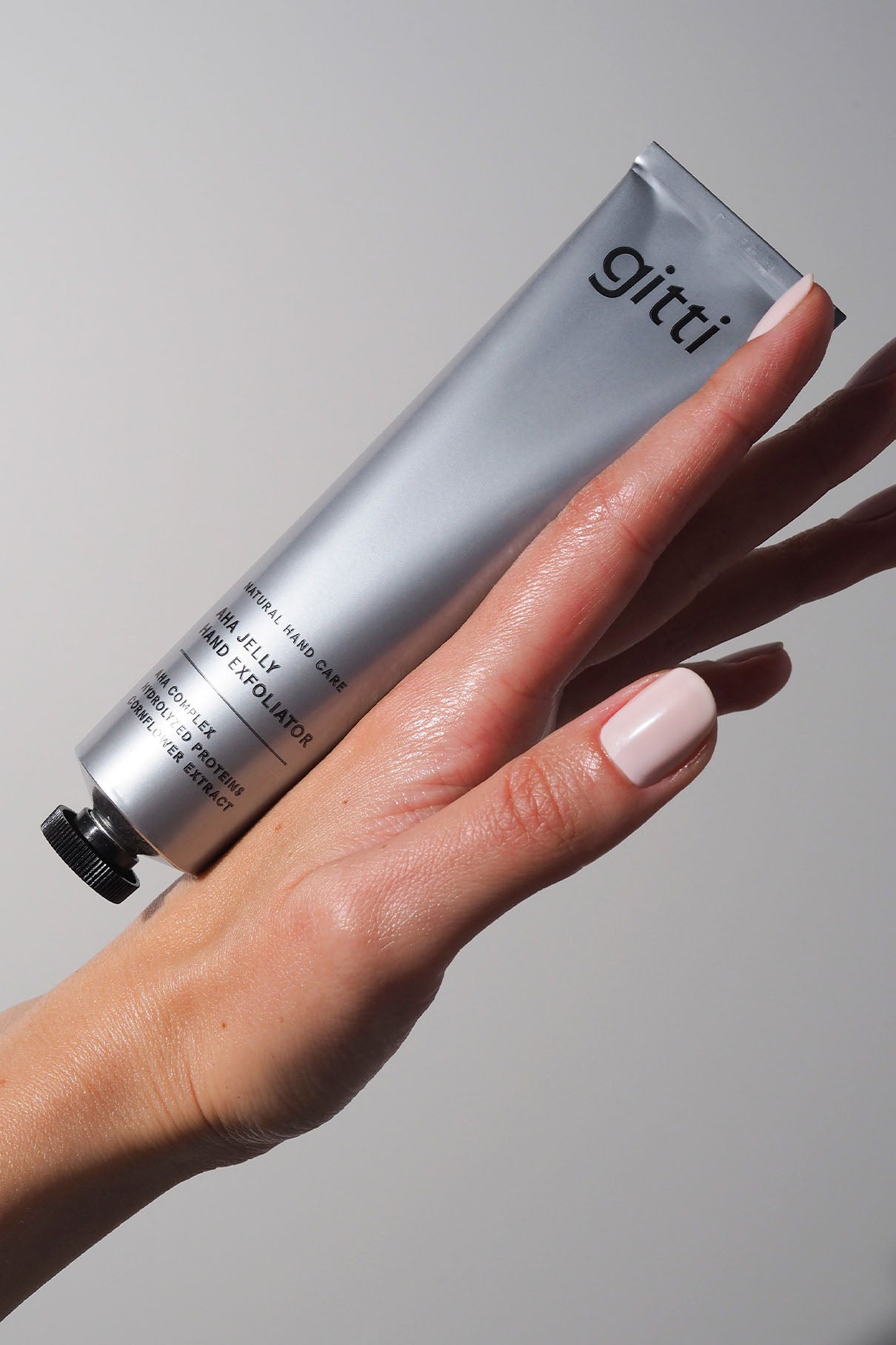 Eine Hand mit ordentlich manikürten Nägeln hält eine silberne Tube mit der Aufschrift gitti Jelly Hand Exfoliator, die ideale Ergänzung zu Ihrem Silky Hands Duo Set, vor einem schlichten Hintergrund.