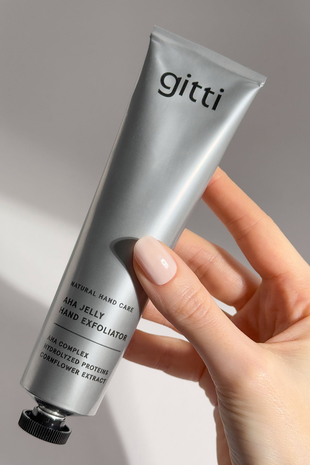 Eine Hand hält eine silberne Tube mit der Aufschrift AHA Jelly Hand Exfoliator mit schwarzer Schrift, die einen AHA-Komplex für sanftes Peeling enthält, vor einem neutralen Hintergrund.