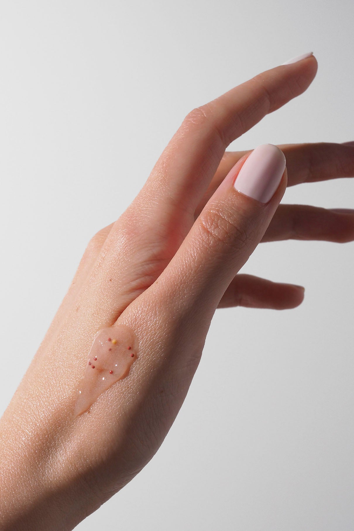 Eine Hand mit blassrosa Nagellack zeigt eine kleine Menge AHA Jelly Hand Exfoliator - ein klares Gel mit winzigen farbigen Kügelchen und einem AHA-Komplex -, das auf den Handrücken aufgetragen wurde.