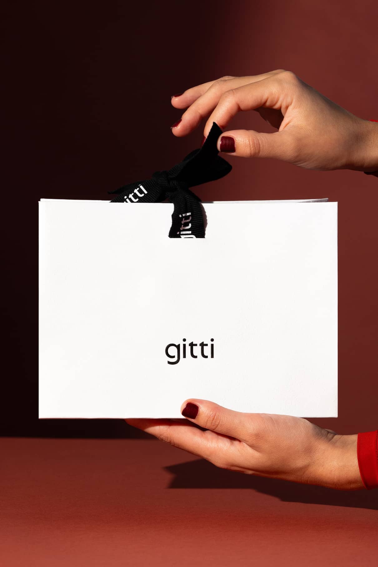 Eine Person hält eine elegante Geschenktüte in der Hand: eine weiße Gitti-Geschenktüte mit einer schwarzen Schleife vor einem rotbraunen Hintergrund.