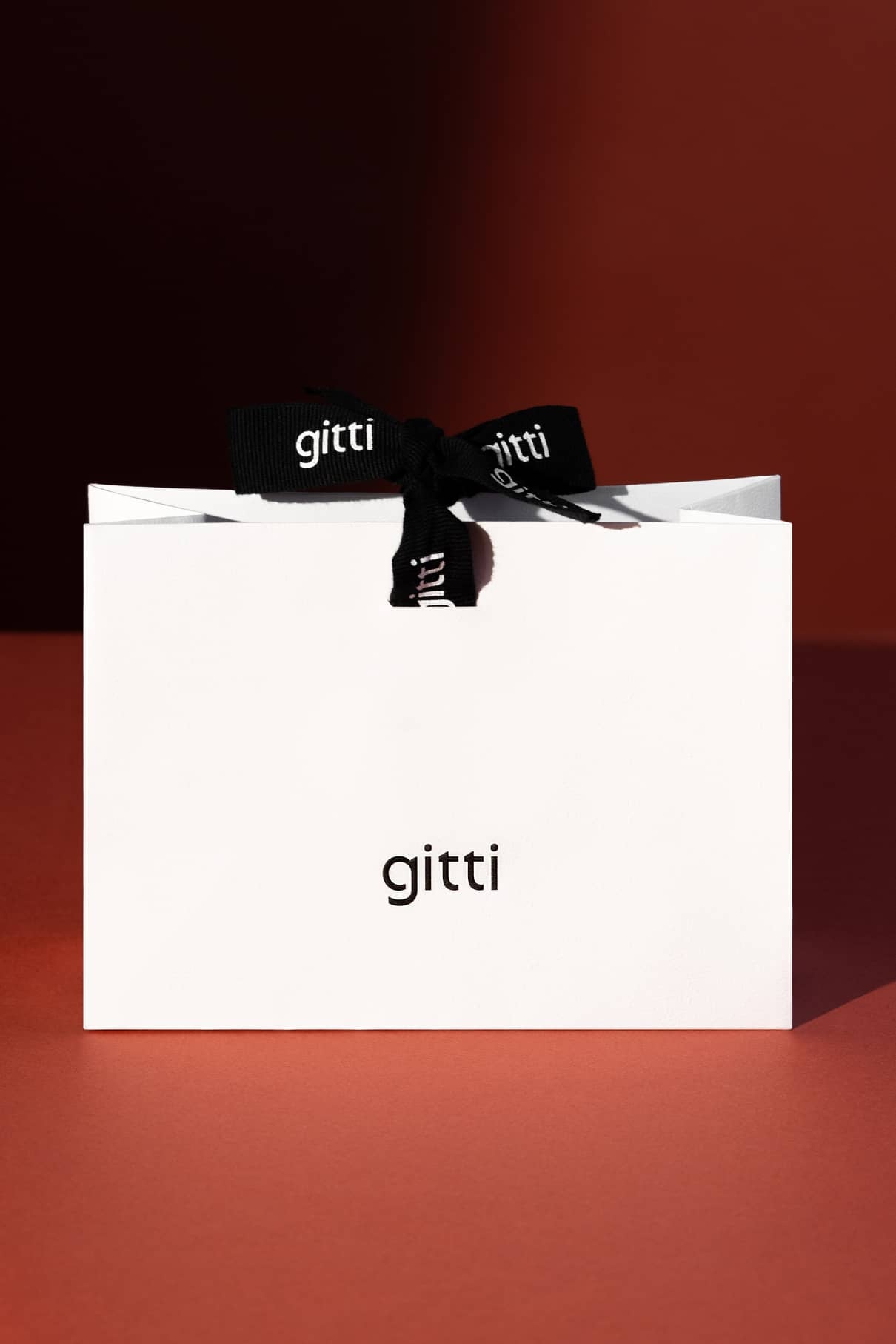 Eine weiße Geschenktüte mit dem Aufdruck "gitti" liegt auf einer rotbraunen Fläche; oben ist ein schwarzes Band mit dem gitti-Logo gebunden.