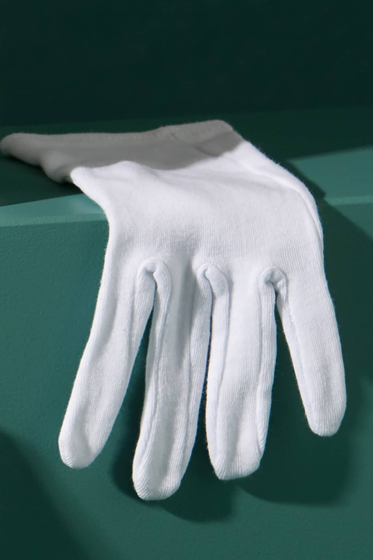 Ein einzelner weißer Baumwoll-Handschuh, OEKO-TEX 100 zertifiziert, liegt über dem Rand einer teal-farbenen Oberfläche mit weichem Schatten.