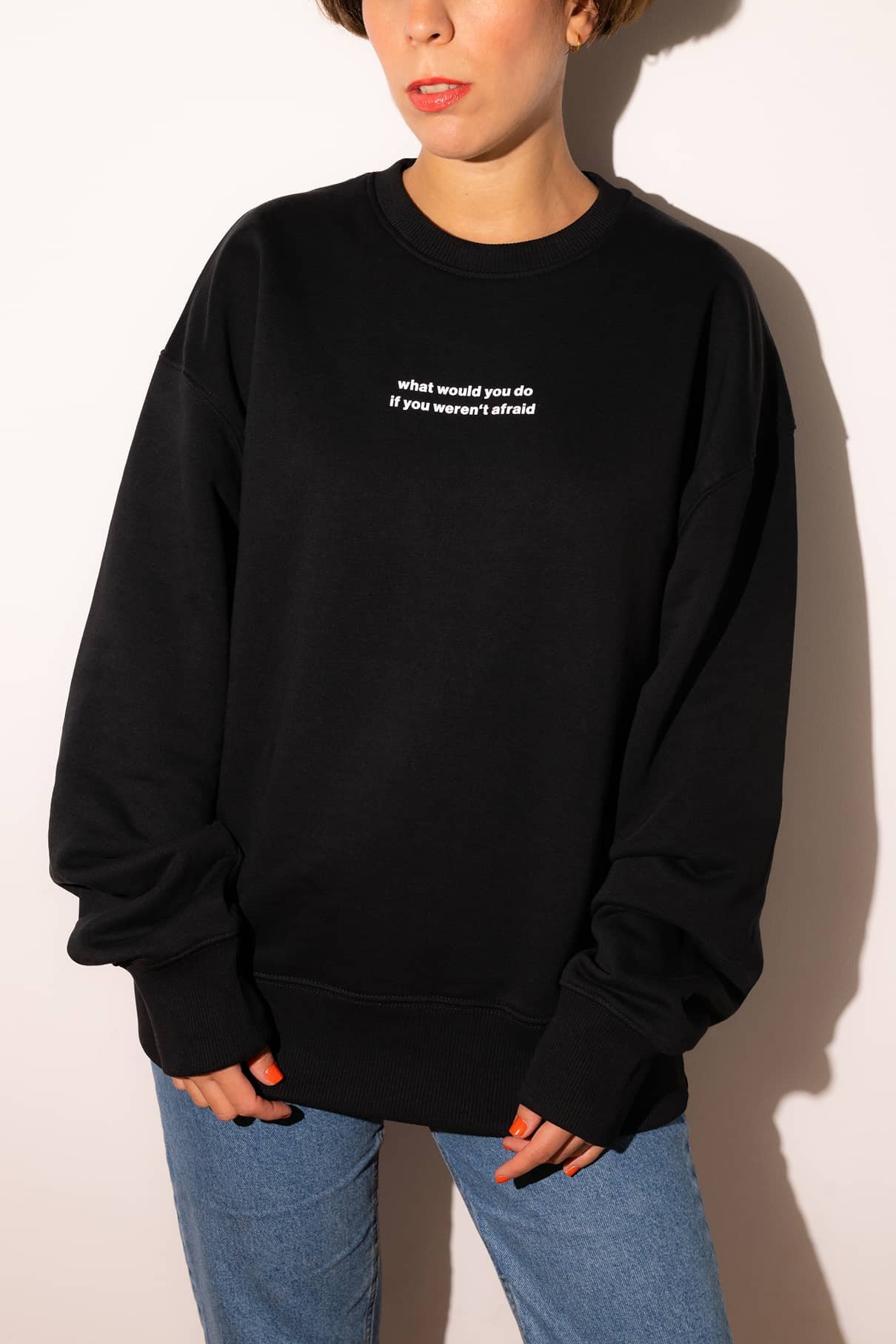 Eine Person steht vor einem schlichten Hintergrund, trägt den schwarzen Gitti-Pullover mit der Aufschrift "Was würdest du tun, wenn du keine Angst hättest?" und blaue Jeans und inspiriert andere, mutig zu sein und ihren Träumen zu folgen.