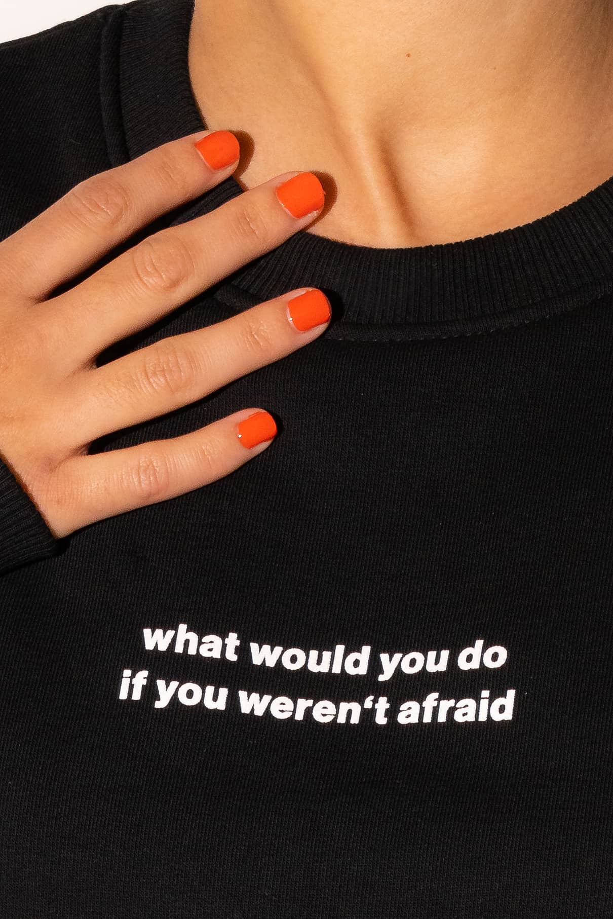 Eine Hand mit orange lackierten Nägeln ruht auf dem schwarzen Gitti-Pullover, auf dem in weißer Schrift steht "what would you do if you werent afraid" - eine stilvolle Erinnerung an "mutig sein" und "Träume verfolgen".
