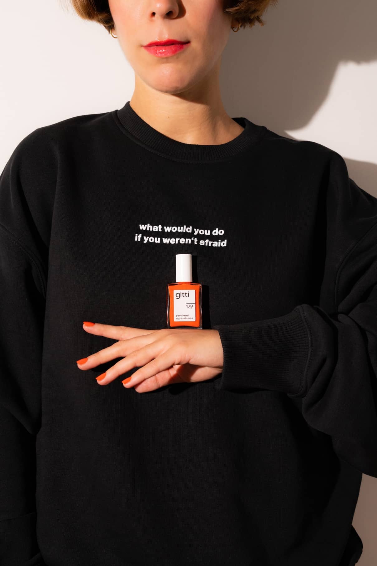 Eine Person in einem schwarzen Gitti-Pullover mit der Aufschrift "Was würdest du tun, wenn du keine Angst hättest" hält orangefarbenen Nagellack in der Hand und inspiriert andere zum Träume verfolgen und mutig sein.