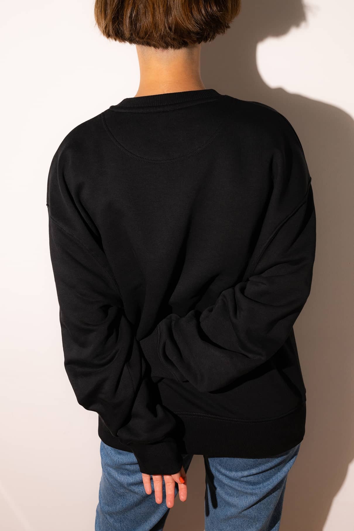 Eine Person mit kurzen braunen Haaren, die einen schwarzen Gitti-Pullover und eine blaue Jeans trägt, steht mit dem Gesicht nach hinten, die Hand auf dem Rücken, bereit, zu mutig zu sein und ihre Träume zu verfolgen.