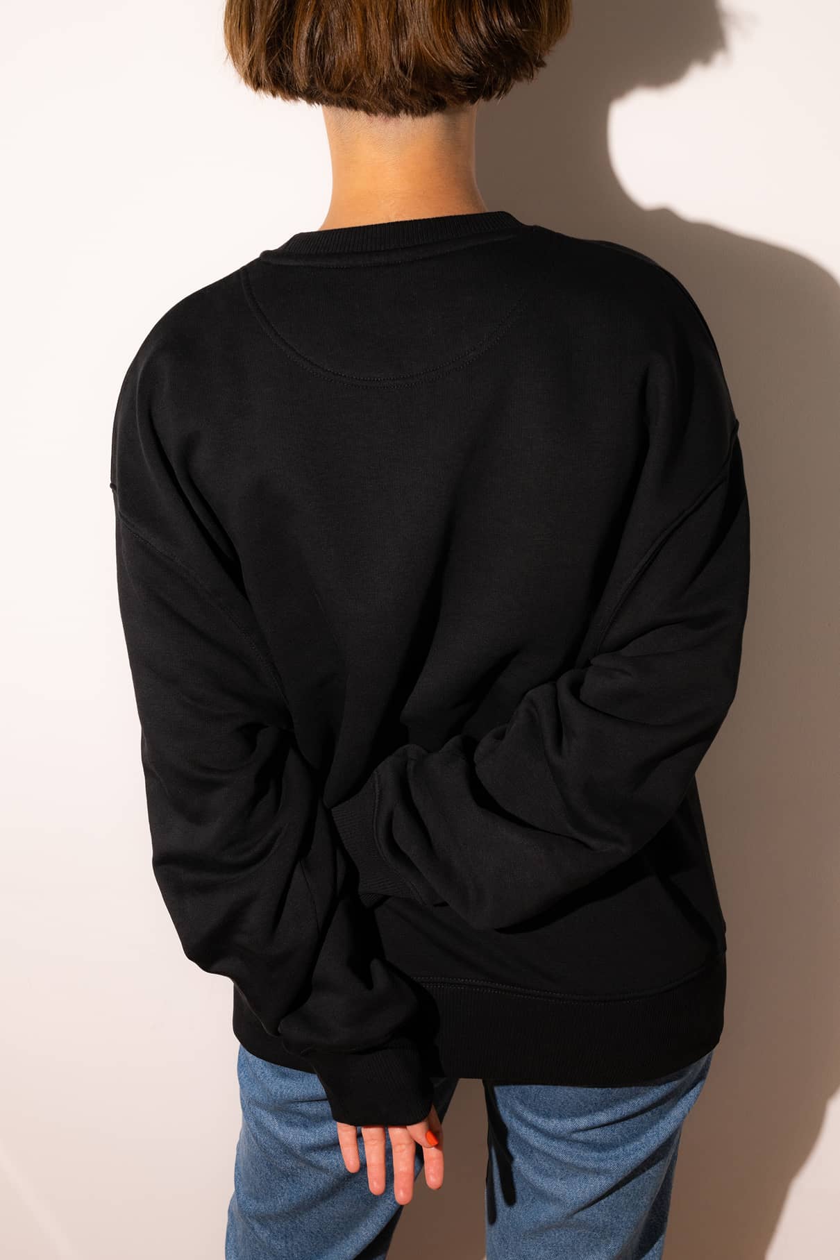 Eine Person mit kurzen braunen Haaren, die einen schwarzen Gitti-Pullover und eine blaue Jeans trägt, steht mit dem Gesicht nach hinten, die Hand auf dem Rücken, bereit, zu mutig zu sein und ihre Träume zu verfolgen.