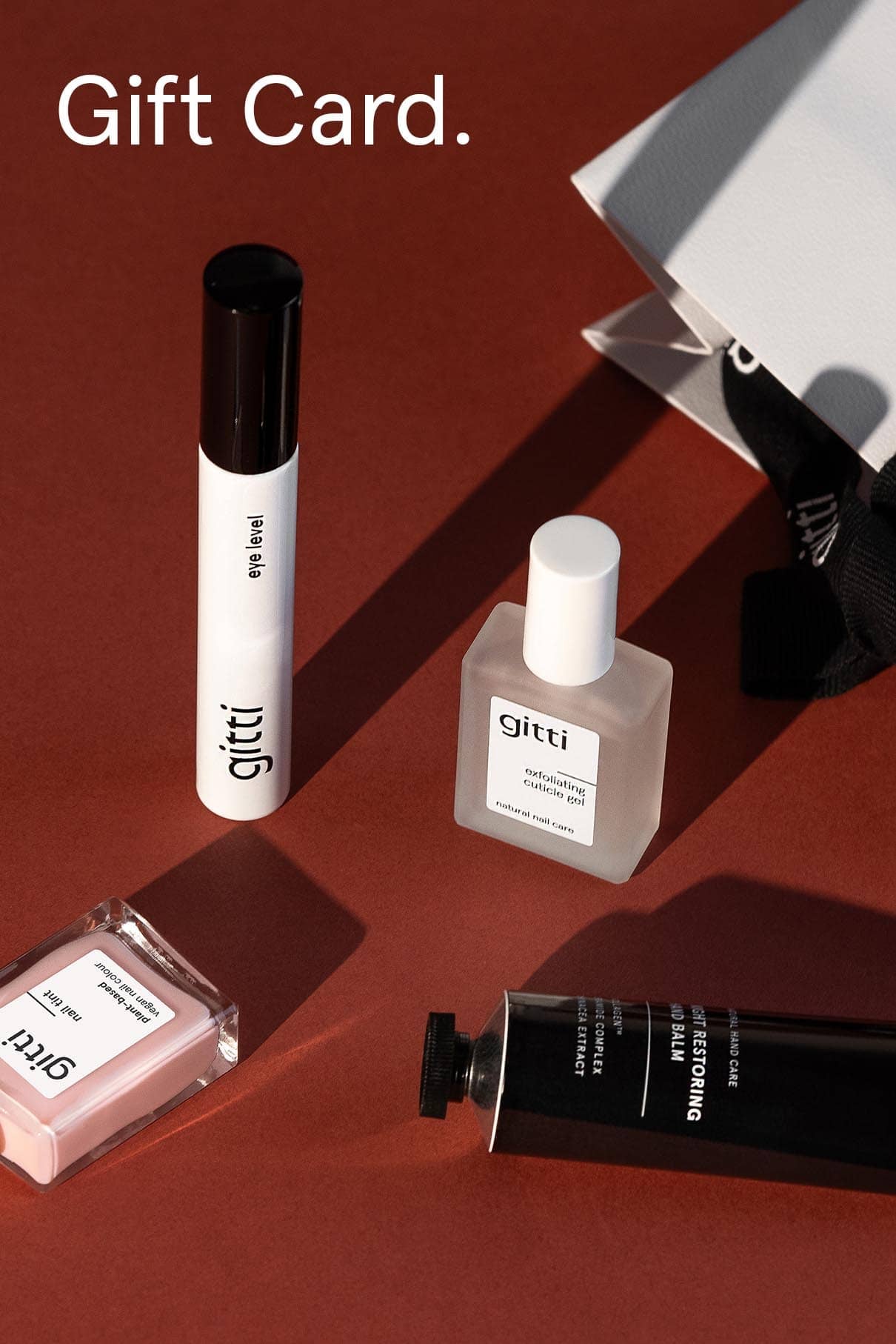 Ein Digitaler Geschenkgutschein mit verschiedenen Gitti-Schönheitsprodukten - Nagellackflasche, Gel-Etikett-Tube, Nagelfeile, schwarze Tube - angeordnet auf einer braunen Fläche neben einer weißen Tasche. Perfekt zum Verschenken Ihrer Gitti-Lieblingsartikel!.