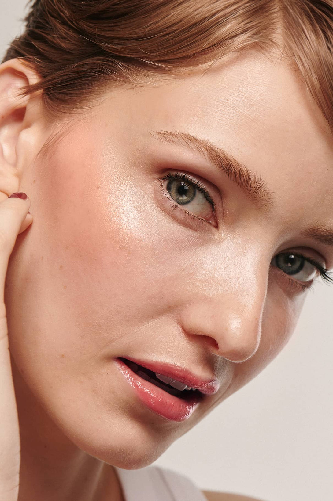 Nahaufnahme einer Frau mit heller Haut, kurzen hellbraunen Haaren und natürlichem Make-up, die sanft ihre Wange berührt, die durch ein subtiles Leuchten von All Glow akzentuiert wird.