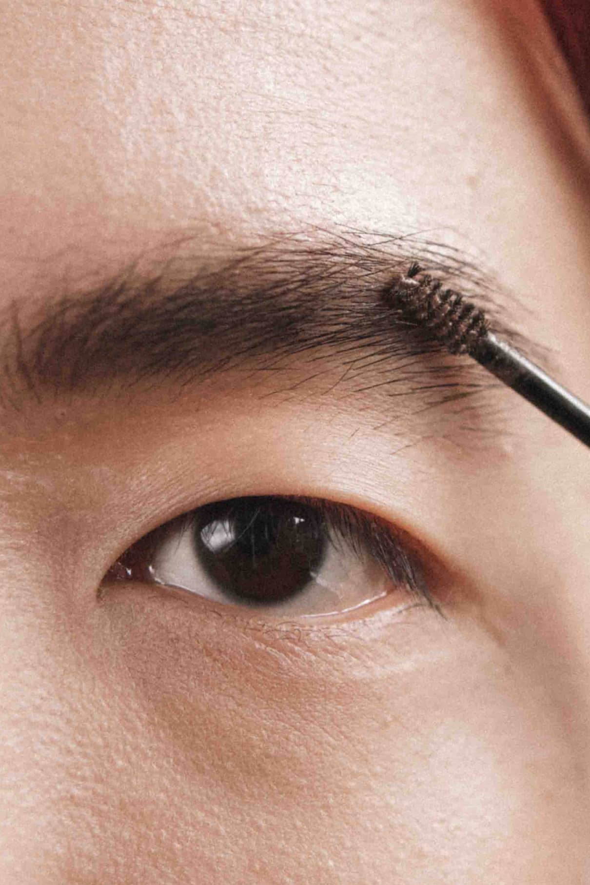 Eine Nahaufnahme des Auges einer Person, die mit einem Spoolie ihre mittelbraunen Augenbrauen pflegt und Berlin Brow 03 Gel aufträgt, um den Look zu perfektionieren.
