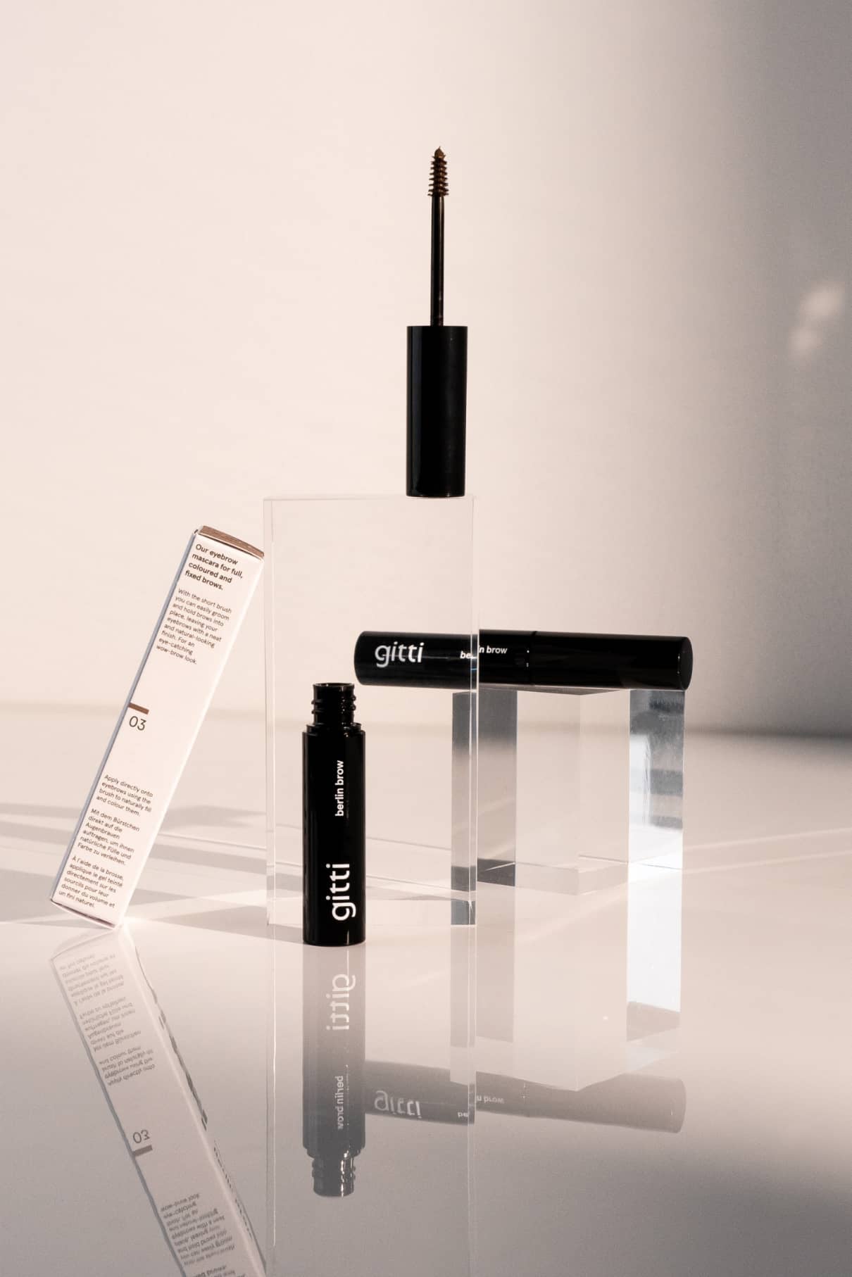 Zwei Tuben Berlin Brow 03, eine davon offen mit sichtbarem Stift, sind mit Verpackung auf klaren Acrylblöcken vor weißem Hintergrund abgebildet.