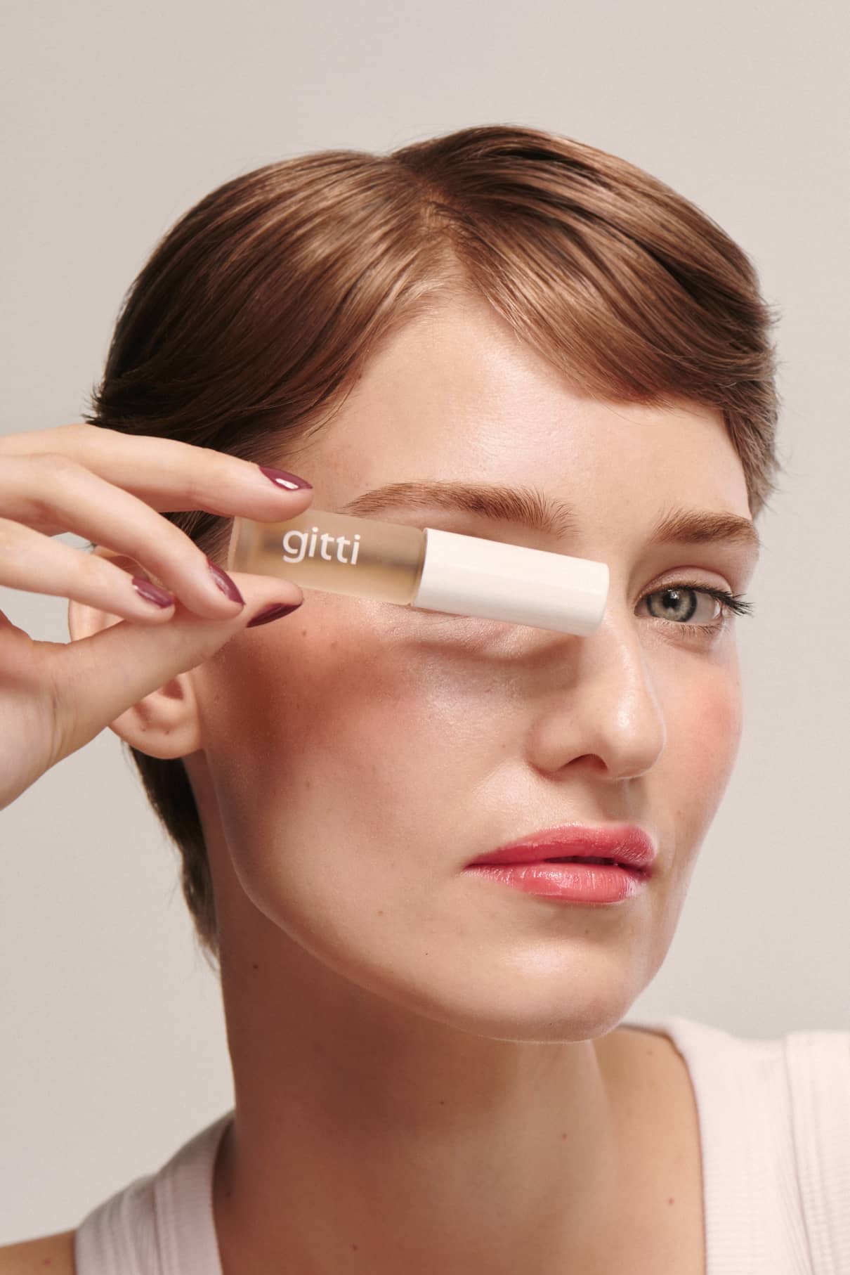 Eine Person mit kurzen braunen Haaren hält ein gitti Brow & Lash Duo vor ihr rechtes Auge, trägt neutrales Make-up und ein weißes Oberteil.